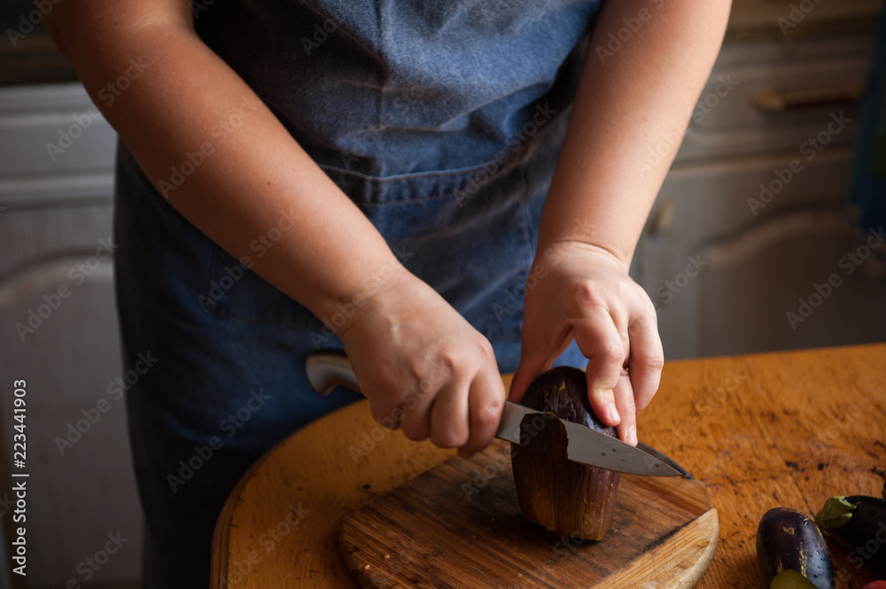 Woman cook in apron and cap cooks in kitchen, cuts eggplants, peppers, onions, garlic on cutting board with large knife. Stir with ladle in saucepan seasoning for canning. Hands close up Woman cook in apron and cap cooks in kitchen, cuts eggplants, peppers, onions, garlic on cutting board with large knife. Stir with ladle in saucepan seasoning for canning. Hands close up