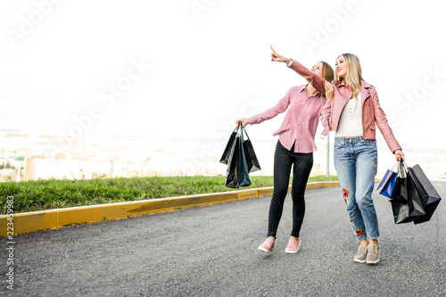 Two young girlfriends walk around the city with shopping bags after a big black Friday sale, pointing the finger to the side