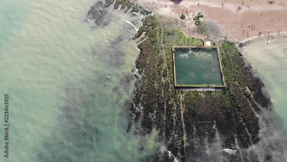 Reverse fly over the man-made paddling pool at low tide in Broadstairs, with clouds and the sun reflecting in the water. Heads up into revealing the Victoria Gardens and Viking Bay, at sunset.
