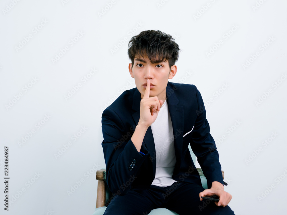 Portrait of handsome Chinese young man in dark blue leisure suit posing against white wall background. Finger on lips and make silent gesture.
