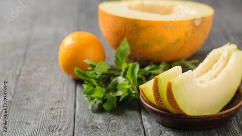 Ripe melon pieces on a clay bowl, banana, mint and orange on a rustic wooden table.