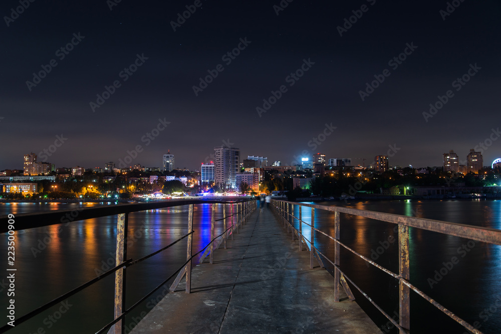 Old long metal pier over the river. Night city landscape