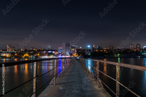 Old long metal pier over the river. Night city landscape