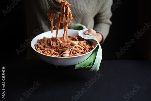 Young woman holding a large white porcelain bowl of whole grain noodles with seafood in one hand, serving pasta with tongs with other hand.Dark background, selective focus photo