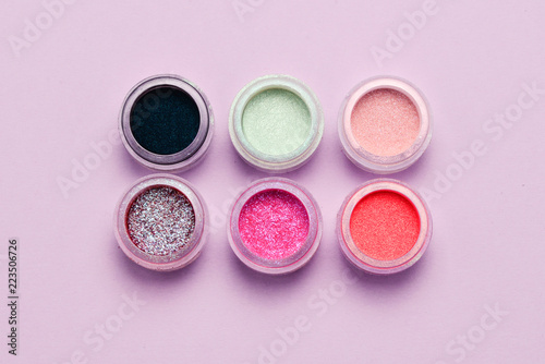 Cosmetics. Makeup. Jars with crumbly bright shadows, glitter. Pink, green, lilac colors on lilac background. Closeup. Space for text or design.