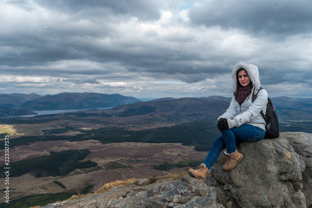 Woman with fluffy white jacket and hiking shoes posing on a mountain with mountains and lakes in the backgroud, on a cloudy day at Nevis Range, Scottish Highlands