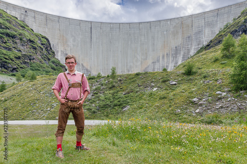 Beautiful handsome young man with glasses in traditional clothing with lederhosen and checkered shirt in summer in front of the Moiry dam near Grimentz, Switzerland
 photo