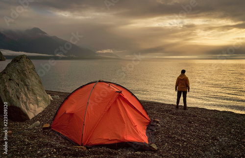 Hiker looking at beautiful lake at sunrise near orange tent.