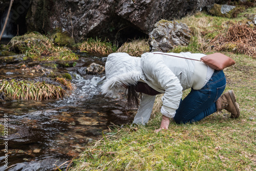 Woman with white puffy jacket and hiking shoes and scarf drinking fresh pure water from the stream at Glencoe Scottish Highlands