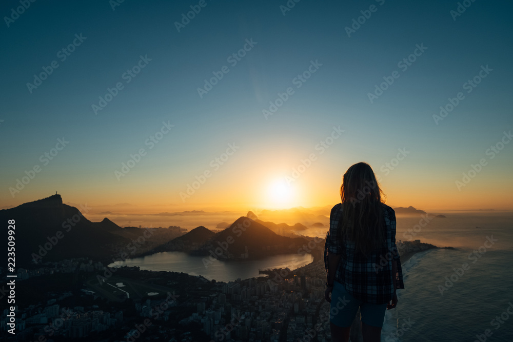 Brazil, Rio de Janeiro. Meeting the dawn on the hill Two brothers. View of the big city, lake, ocean, hills, mountains. Orange-blue colors Silhouette of a girl who looks at the city and sun.