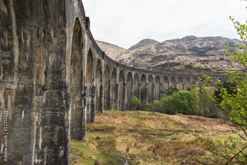 Old train railway with arched bridge surrounded by trees with hills in view, Glennfinnan Viaduct, Scottish Highlands.