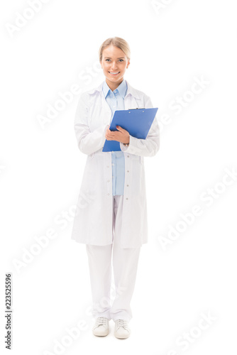 smiling young female doctor writing in clipboard and looking at camera isolated on white