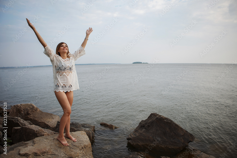 Young beautiful woman in a white tunic is standing on gray granite stones on the beach. Young beautiful woman in a white tunic is standing on gray granite stones on the beach.