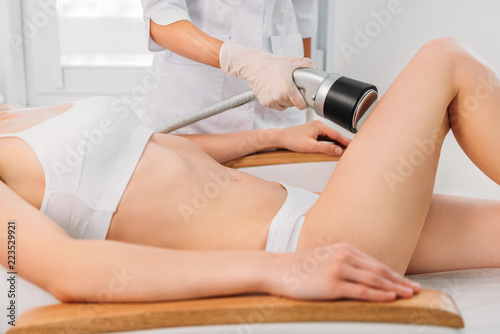 partial view of cosmetologist making electric massage on leg to woman in white underwear at spa salon