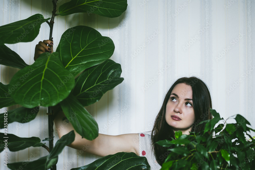 Fototapeta premium Pretty brunette girl sitting near flowerpot on the floor of the living room and looking in the distance. Cozy leisure atmosphere at home.