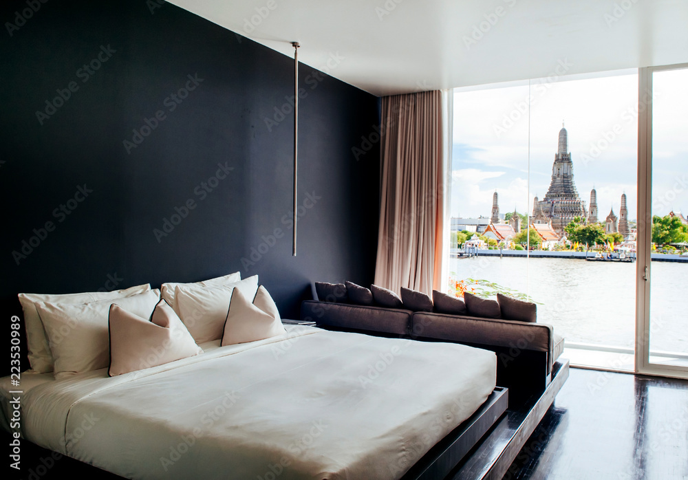 Fototapeta premium Bedroom with black wall white bed, pillows couch, lamp. Bay window with Wat Arun Temple view. Bangkok boutique hotel modern urban interior.
