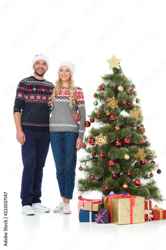 beautiful happy couple in santa hats standing near christmas tree with gifts, isolated on white beautiful happy couple in santa hats standing near christmas tree with gifts, isolated on white