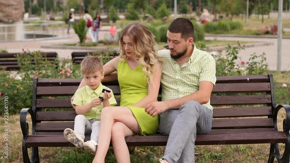 Happy young family with his son resting in the Park in the summer on a bench. A beautiful woman with her husband and a child sitting on a bench in the background of the urban landscape. Happy young family with his son resting in the Park in the summer on a bench. A beautiful woman with her husband and a child sitting on a bench in the background of the urban landscape.