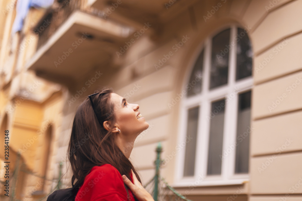happy tourist woman walking on the street with nice architecture. brunette girl is looking at something. wearing red hoodie and with packed black bag