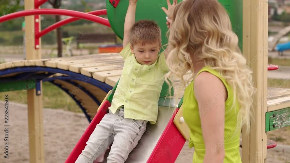 A beautiful young mother helps her son to leave the hill on the playground. Happy mother and son playing in the playground in the summer park. A beautiful young mother helps her son to leave the hill on the playground. Happy mother and son playing in the playground in the summer park.