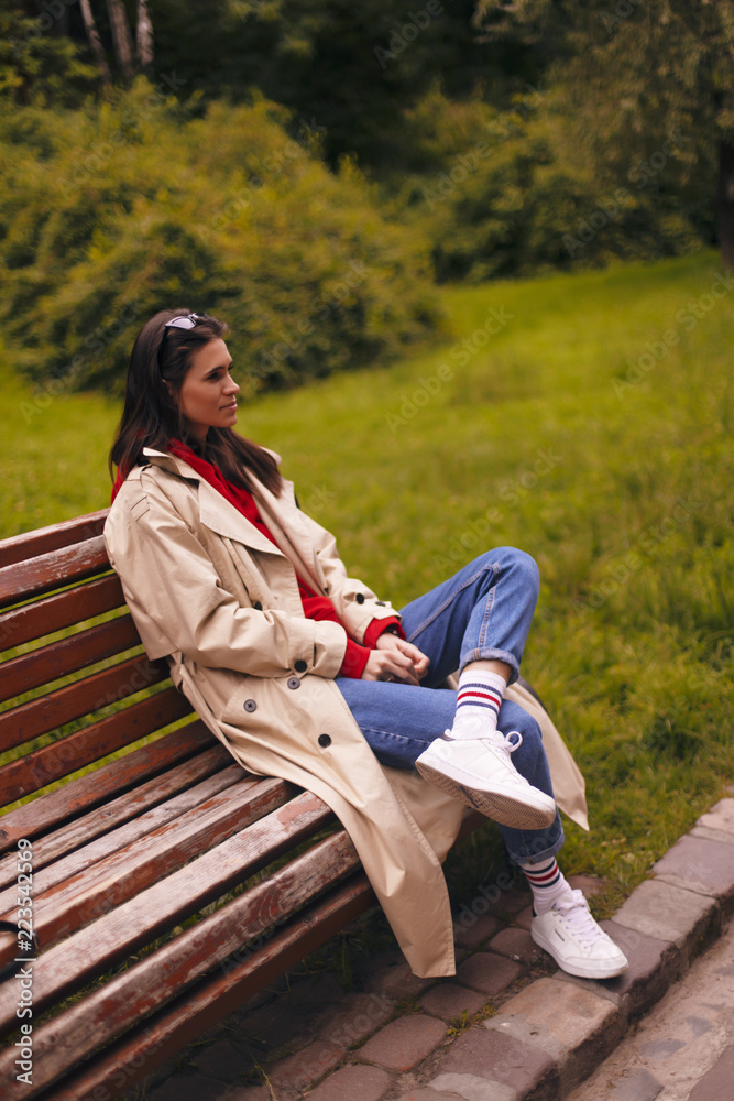 Portrait of woman in full length wearing beige trench coat, red hoodie, blue jeans and white sneakers while walking in park. Portrait of woman in full length wearing beige trench coat, red hoodie, blue jeans and white sneakers while walking in park.