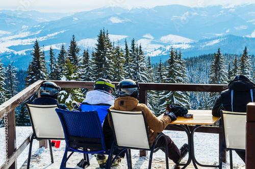 skiers sit on chairs near a restaurant on top of a mountain. People in ski suits rest and drink
