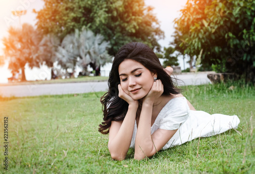 In selective focus of the beauty lady is wearing white dress,laying down on green grass ground floor,closed her eyes,with smile and happy face,relax time,in a park,warm light tone,blurry light around