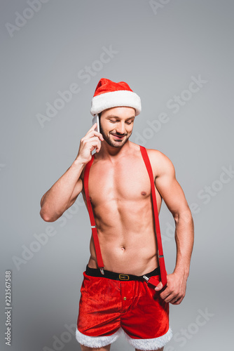 smiling shirtless muscular man in christmas hat talking on smartphone isolated on grey background