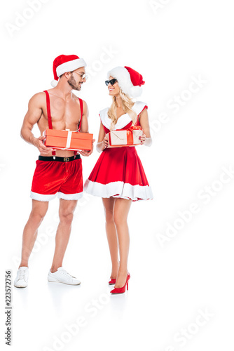 young couple in christmas hats and sunglasses holding gift boxes and looking at each other isolated on white background