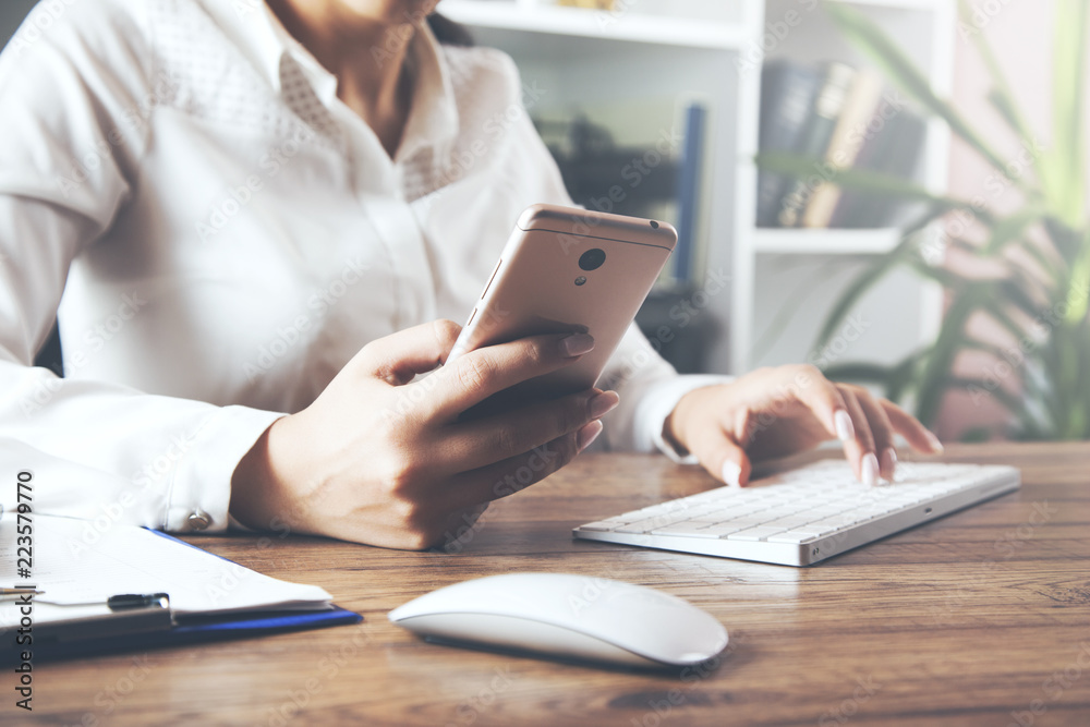 Fototapeta premium Woman one hand holding a phone while the other hand typing on laptop keyboard