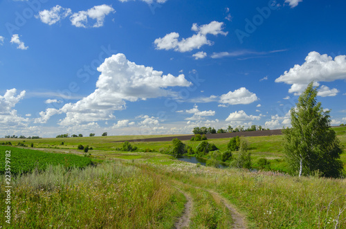 Sunny summer landscape with ground country road,fields,green hills and beautiful clouds in blue sky.