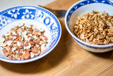 A blue bowl of chopped almonds and a blue bowl of chopped peanuts on a bamboo cutting board.