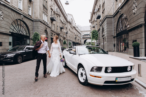 A newlyweds trip in the car. The groom invites the bride in a white cabriolet.