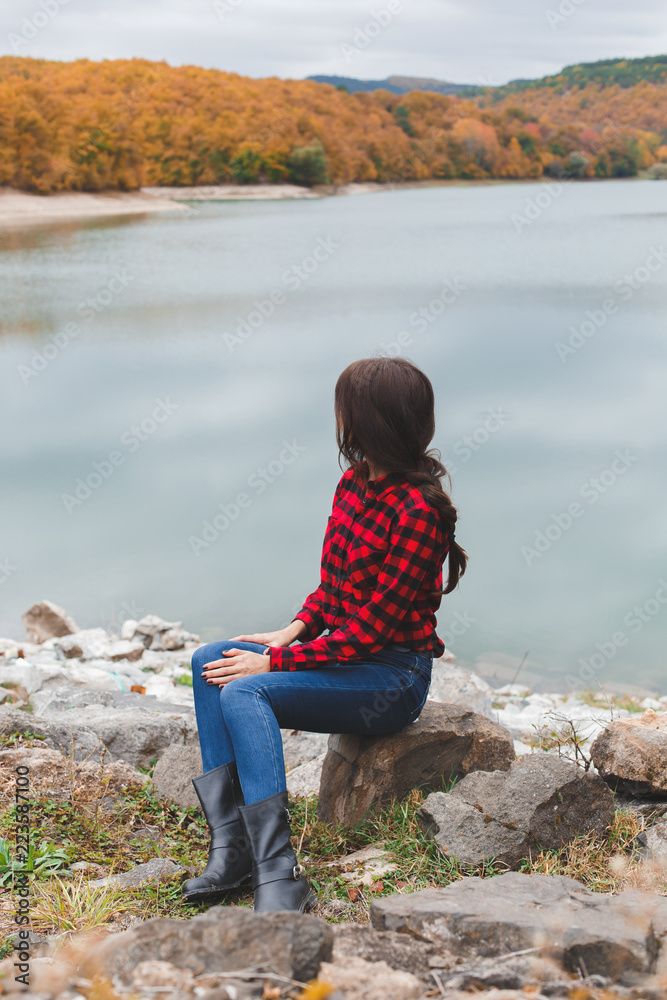 Young woman wearing red plaid shirt sitting alone near the lake in autumn. Weekend outdoors, cloudy cold weather, fall colors. Back view.