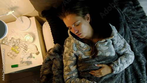 Top view of young sick woman with fever lying in bed at night and coughing. Medicines, thermometer and glass of water on the table.