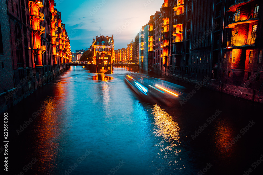 A colorfully illuminated boat cruising on the Wandrahmsfleet at night. The Warehouse District in Hamburg, Germany. The district is located within the HafenCity quarter. Long exposure