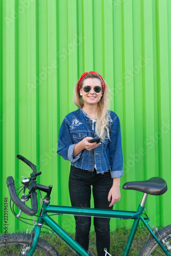 Stylish hipist girl stands with a bike and a smartphone in her hands against the background of a green wall, looks at the camera and smiles. Sports and Technology.