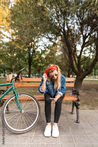 Stylish girl sitting on the bench in the park, with a bike and using a smartphone. A woman uses an internet connection on a smartphone while walking in the park.