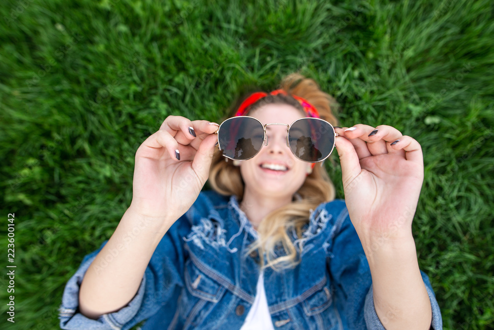 Happy girl lying on green grass holding sunglasses in her hands, looking at camera and smiling, top view. Focus on glasses. Copyspace Happy girl lying on green grass holding sunglasses in her hands, looking at camera and smiling, top view. Focus on glasses. Copyspace