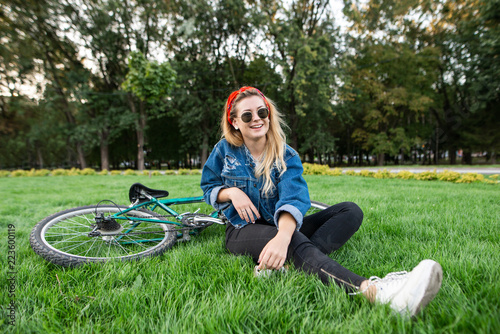 Smiling student girl in stylish clothes sitting on a green lawn in a park with a bike and relaxing. Happy attractive woman in sunglasses in a park with a bike. Outdoor recreation © bodnarphoto