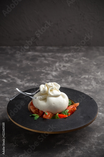 Whole tied Italian cheese burrata on small wooden plate served with fresh tomatoes and basil on dark textured background
