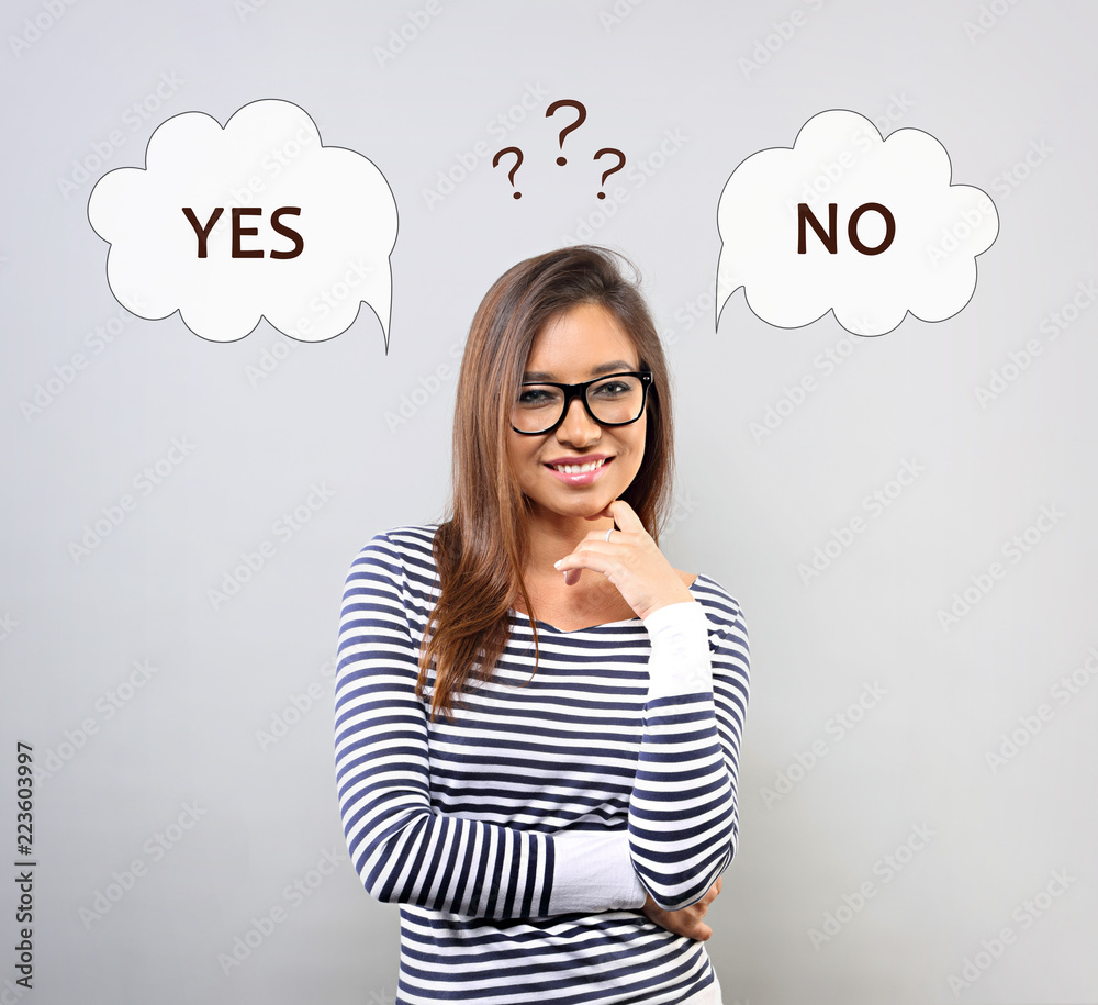 Thinking smiling young latina woman in glasses with two empty choice bubble above with yes and no answers and questions above the head on blue background Thinking smiling young latina woman in glasses with two empty choice bubble above with yes and no answers and questions above the head on blue background