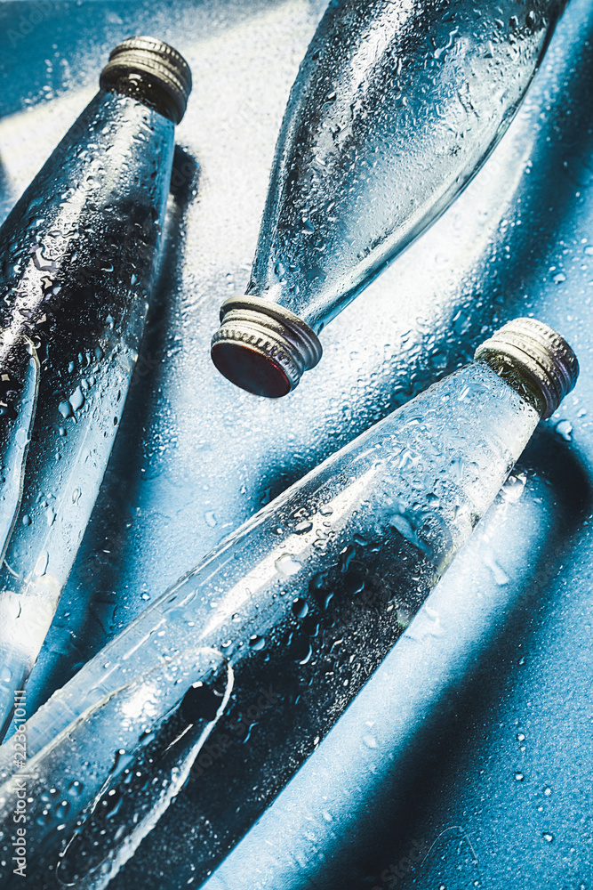 A glass bottle of water on blue background splashes drops of water on top. Quench thirst. Background of water drops