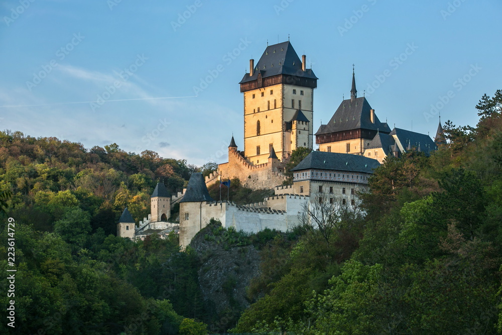 Famous historical medieval castle Karlstejn built by emperor Charles IV., lit by evening sunlight, standing on a green hill covered with trees, Czech Republic, Europe, blue sky