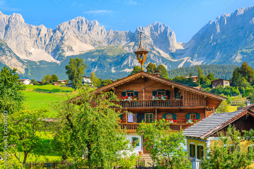 Typical wooden alpine house against Alps mountains background on green meadow in Going am Wilden Kaiser village on sunny summer day, Tyrol, Austria