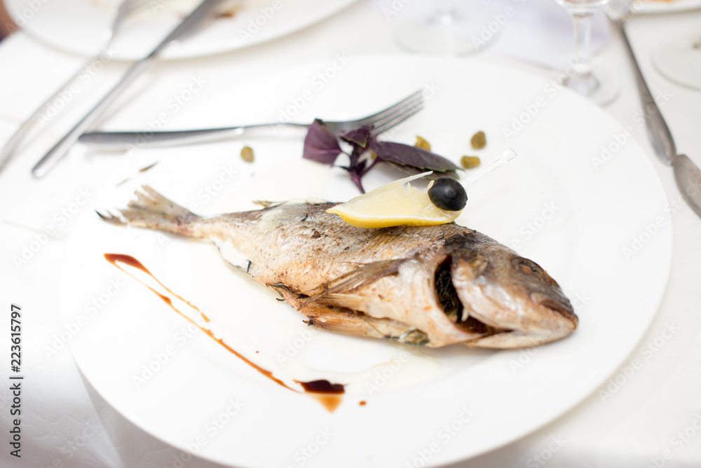 Grilled dorado fish on the white plate with tomatoes and slices of lemon with balsamic sauce, on a white table. Grilled dorado fish on the white plate with tomatoes and slices of lemon with balsamic sauce, on a white table.