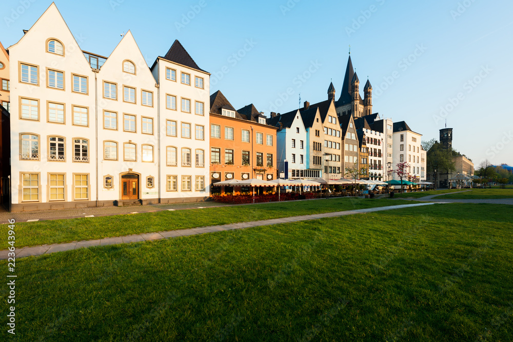 Houses and park in Cologne, Germany. Many of them are colourful, they are facing a public park with green grass and some trees. There is a Cologne bell tower on background. 