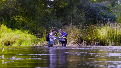 Two scientists ecologists in high rubber boots standing in the water of the forest river. Woman is getting water sampels, man is holding toolbox. Field work photo