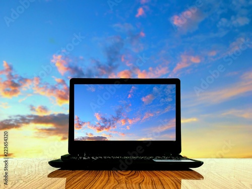 Black laptop and sunrise sky picture in monitor on wooden table with blur sunset sky background in look at the world wild through laptop screen  photo