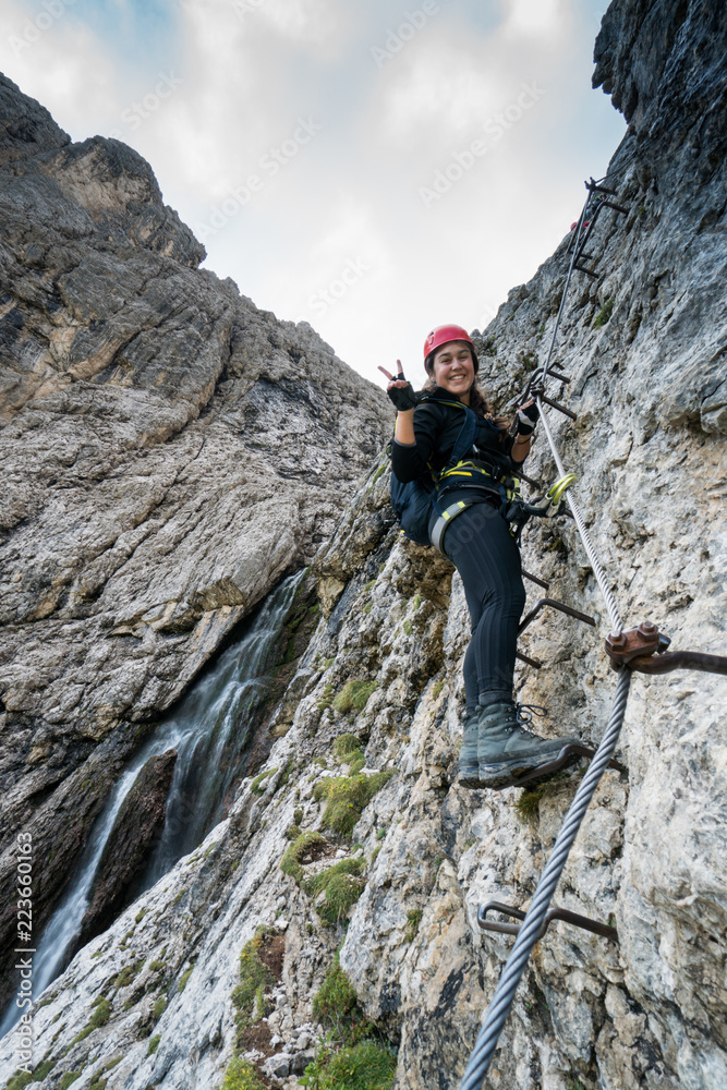 young attractive female mountain climber on a difficult Via Ferrata in the Dolomites in Alta Badia in the South Tyrol in Italy making a peace sign and smiling